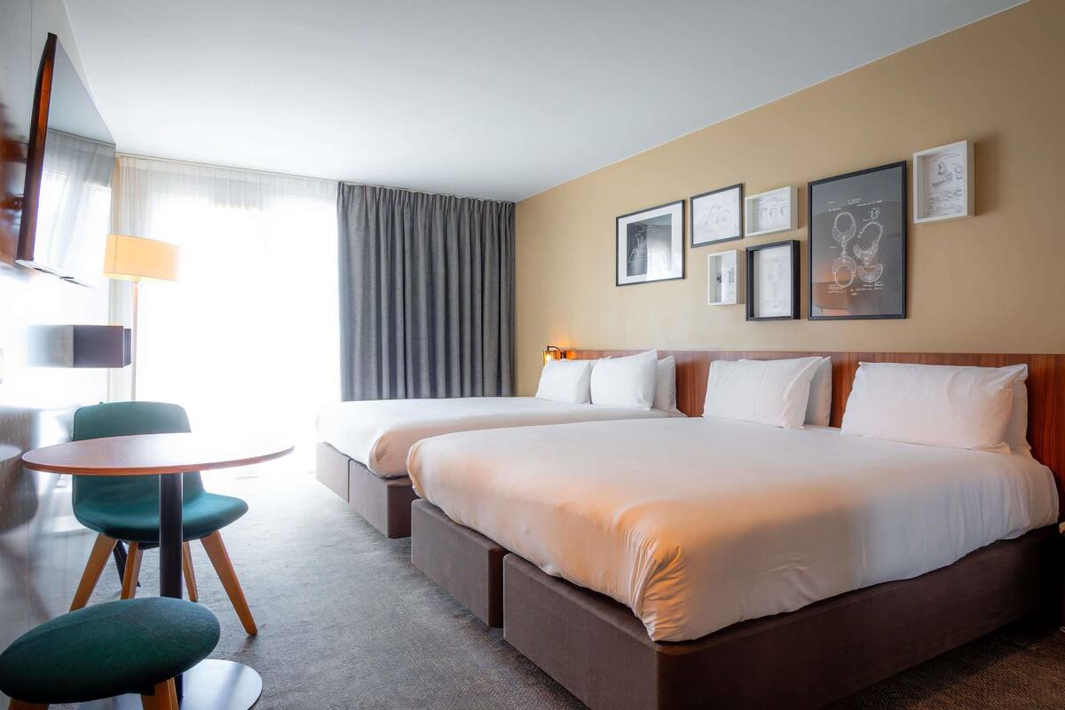 A hotel room with two beds, a small round table, a chair, and a lamp.