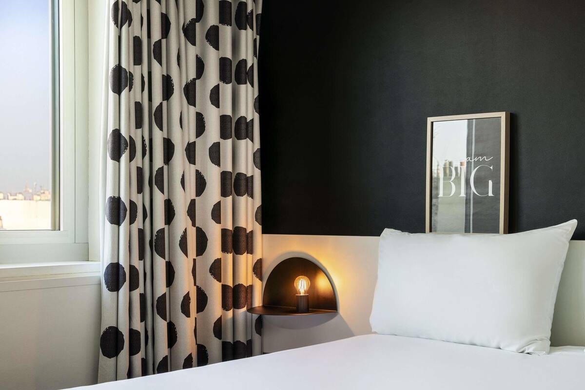Habitación de hotel con cama, luz empotrada en la pared, cita enmarcada y vista a la ciudad desde la ventana.