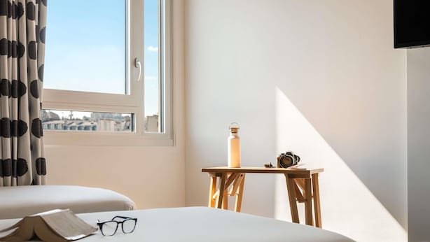 A hotel room with a bed, a small table with a water bottle and camera, and a window with curtains. - ibis Styles Paris Gare de l'Est Château Landon (Paris)