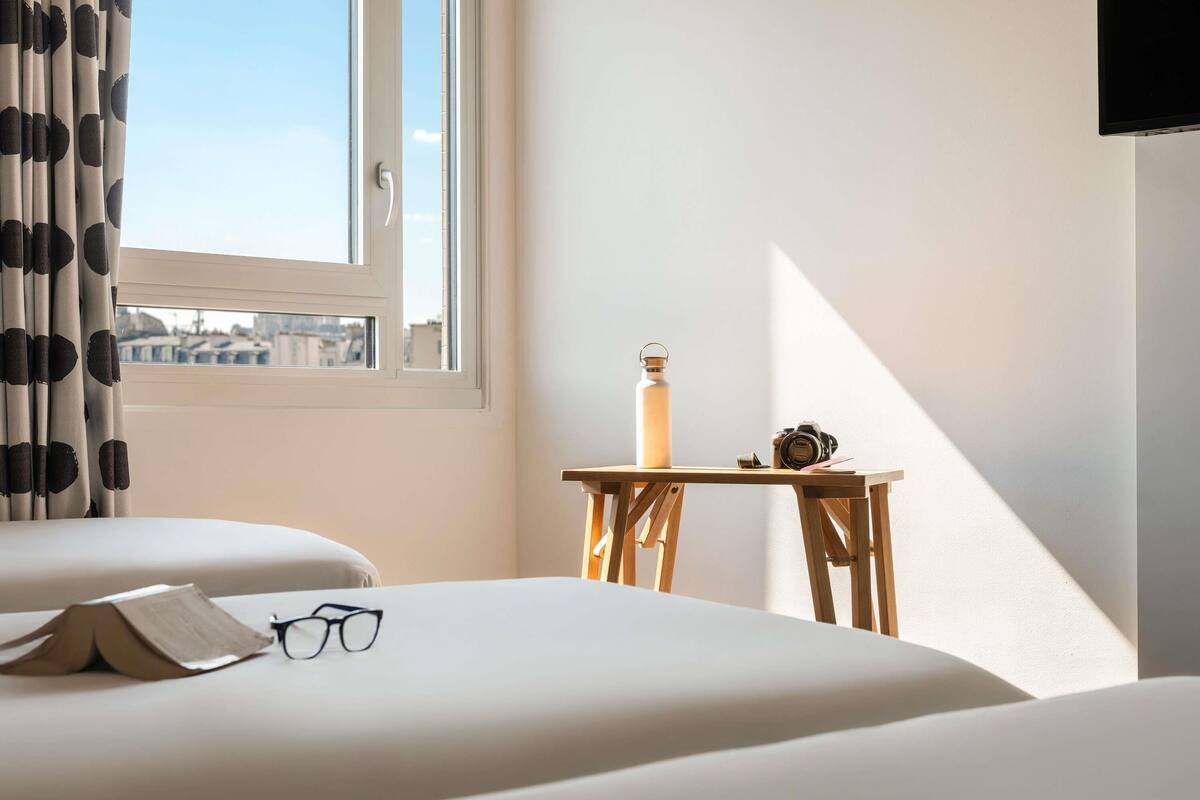 Una habitación de hotel con cama, una mesita con una botella de agua y una cámara, y una ventana con cortinas.