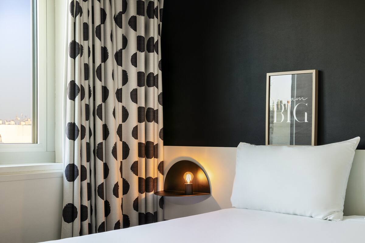 Una habitación de hotel con una cama, una mesita de noche con lámpara, una ventana con cortinas y un cuadro en la pared.