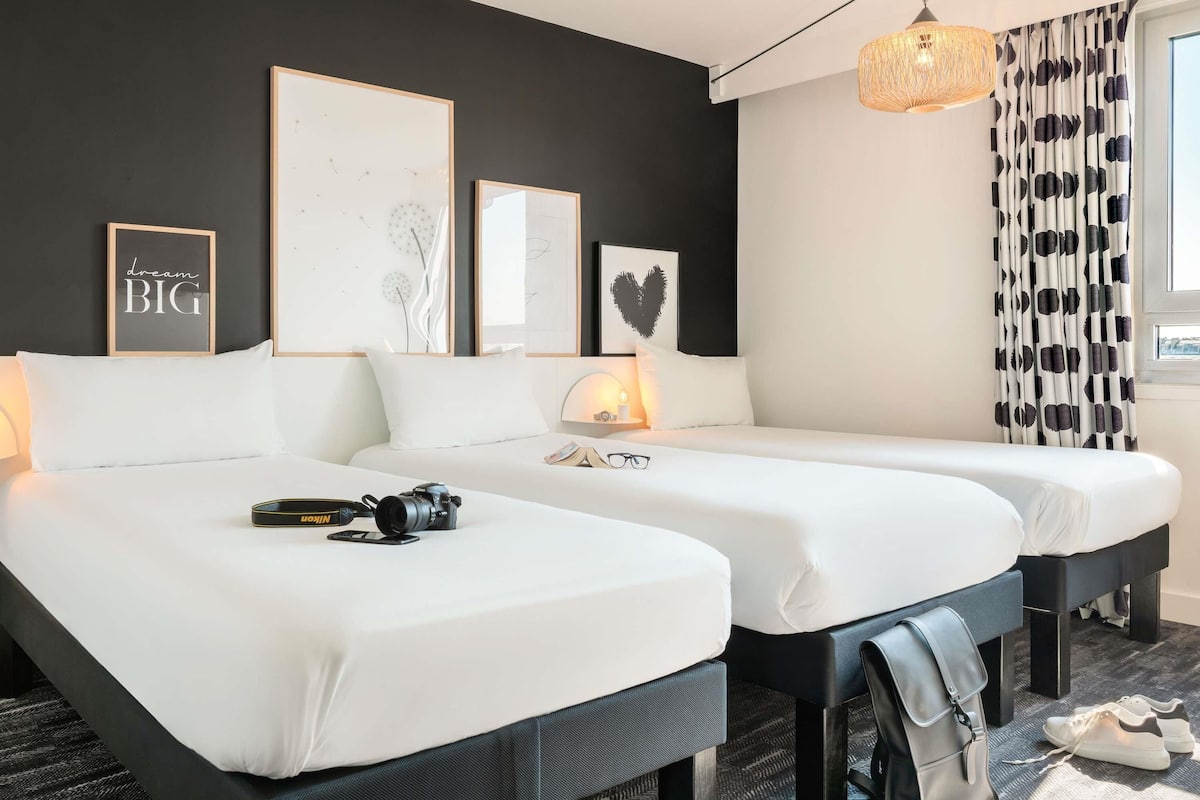 Hotellihuone, jossa on kaksi sänkyä, työpöytä, lamppu ja kamera, joka on sijoitettu toisen sängyn päälle.