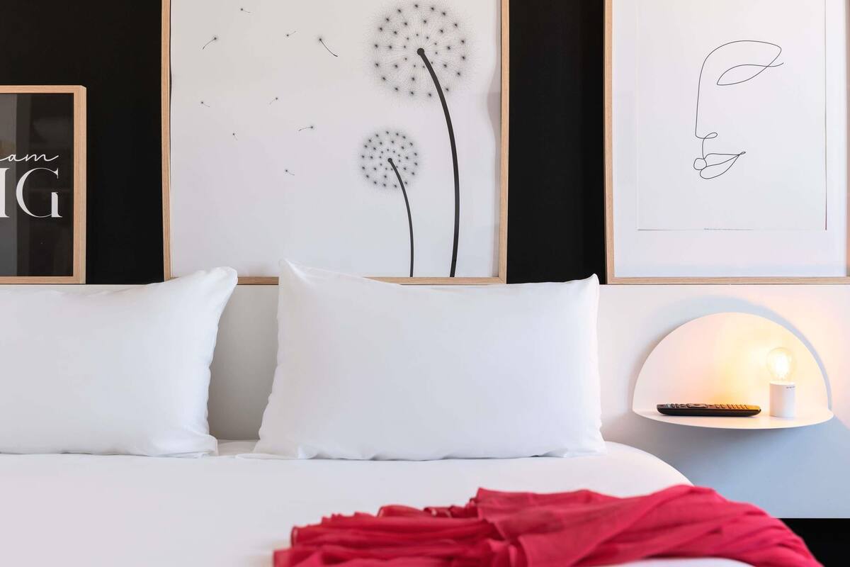 Una cama bien hecha con ropa de cama blanca y una manta roja, una lámpara de pared y cuadros en la pared.
