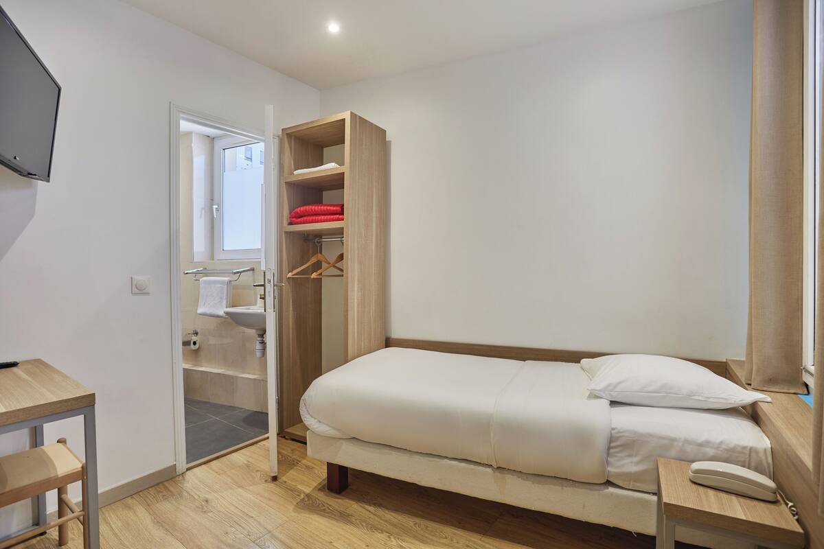 A bedroom with a bed, a wooden wardrobe, a bedside table, and a TV mounted on the wall.