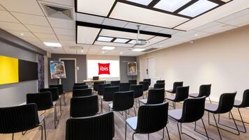 A conference room with a projector screen displaying 'ibis,' black chairs arranged in rows, a wooden floor, and a ceiling with skylights.