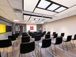 Un salón de conferencias con una pantalla de proyector mostrando 'ibis', sillas negras dispuestas en filas, suelo de madera y un techo con tragaluces.