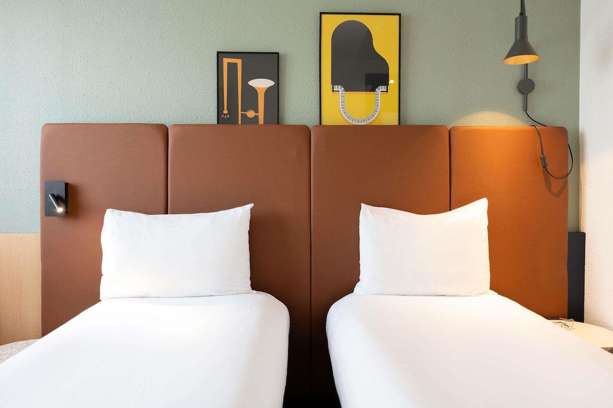 Une chambre dâhĂŽtel avec deux lits, une tĂȘte de lit ornĂ©e dâune Ćuvre dâart, une lampe fixĂ©e au mur et une table de chevet.