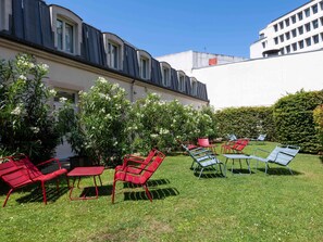 Un patio con mesas al aire libre, un edificio con techo curvo y un seto.