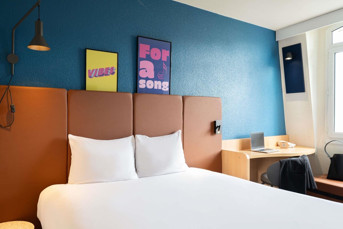 Habitación de hotel con cama, escritorio, silla y una mesita. Se observan dos adornos en la pared y una lámpara.