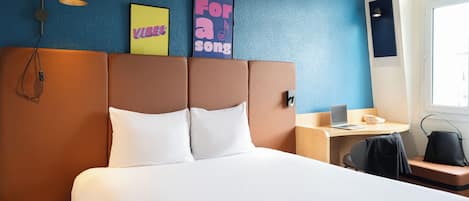 A hotel room with a large bed, a desk with a laptop, a chair, and a small round table. There are two wall art pieces and a lamp.
