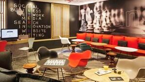 Un área de estar moderna con diversas opciones de asientos, incluyendo sofás y sillones, dispuestos alrededor de mesas y un espacio central con un mural grande de un tablero de ajedrez.