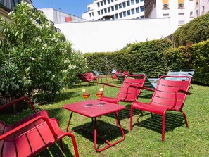 Un patio con sillas y mesas de metal rojo, rodeado de vegetación y edificios.