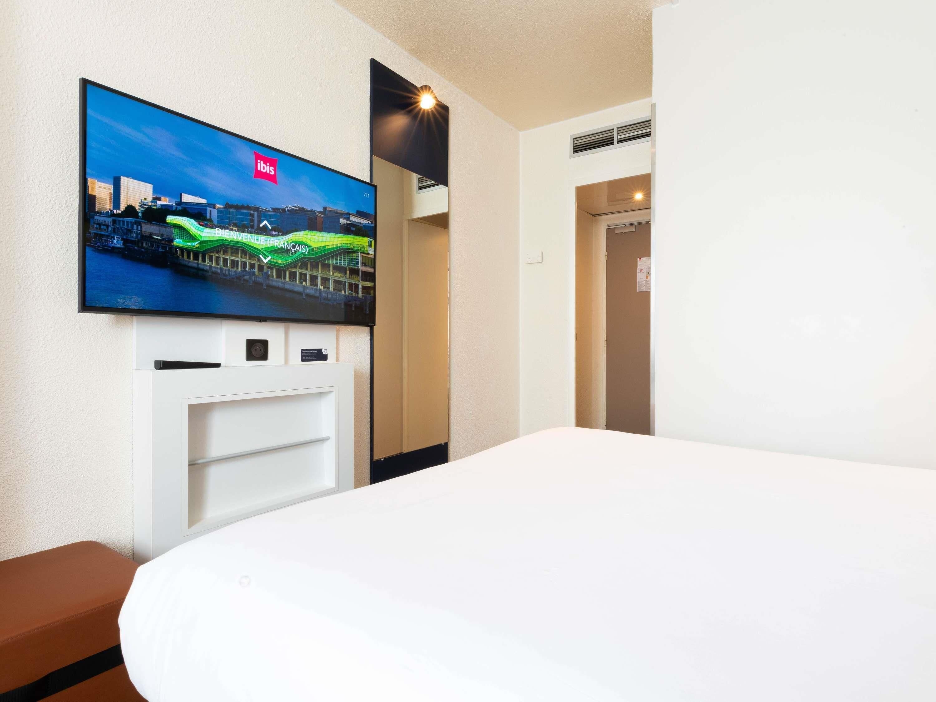 A hotel room with a television mounted on the wall displaying a cityscape, a bed, and a white bedside table.