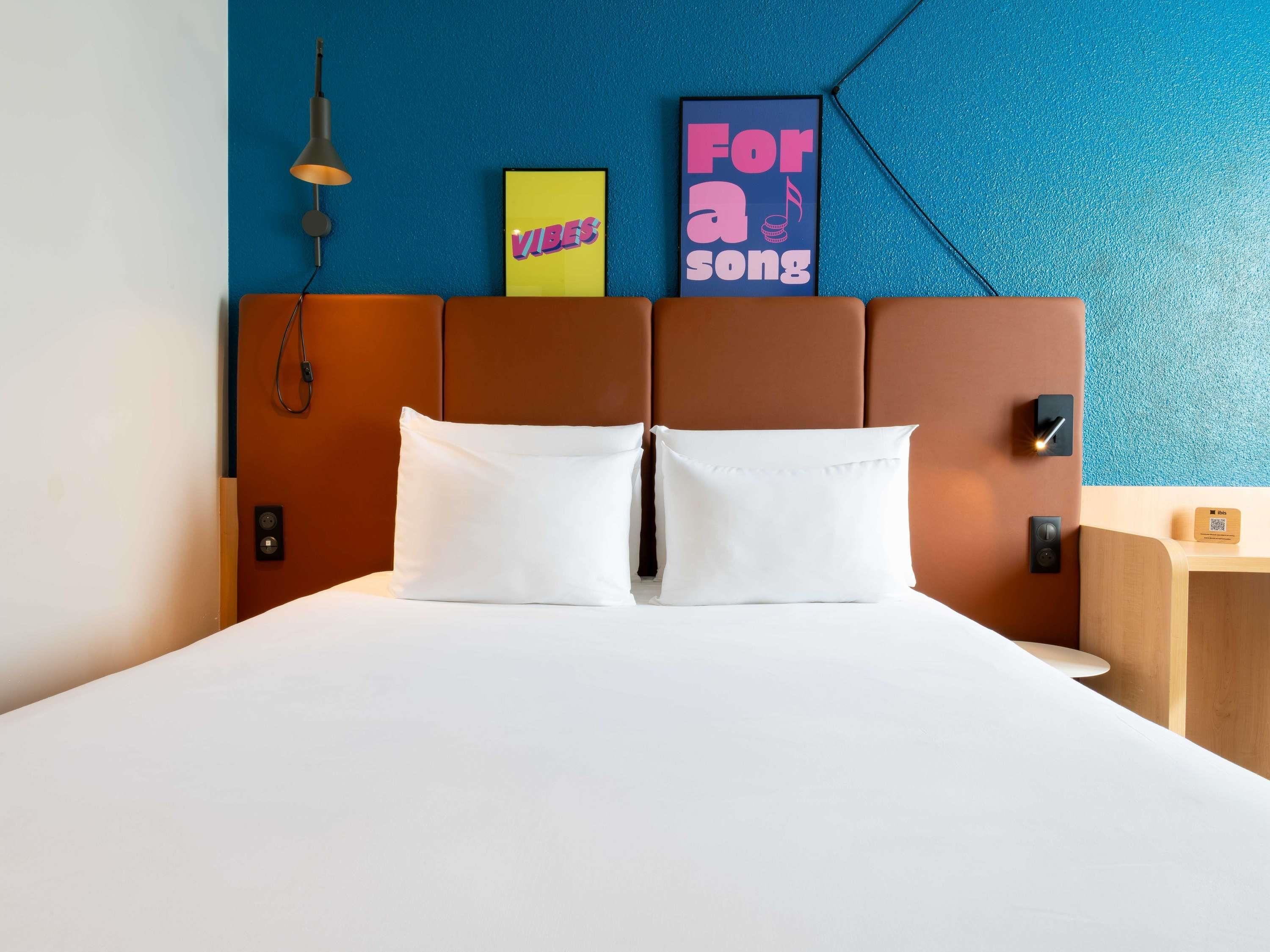 A hotel room with a bed, a headboard, a wall-mounted lamp, and a framed poster.