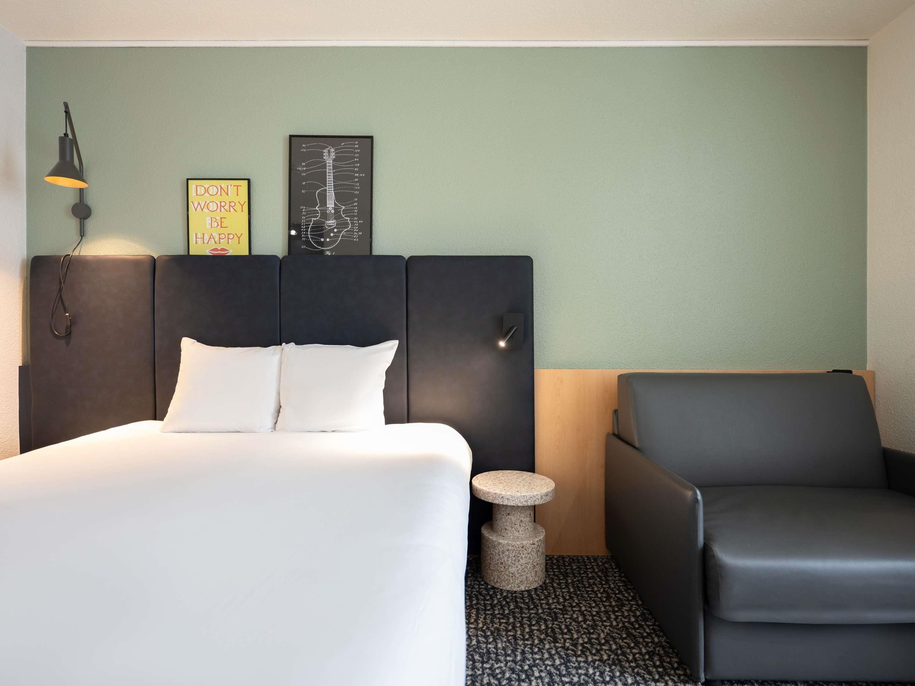 Habitación de hotel con cama, una silla, una lámpara de pared, un diagrama de guitarra enmarcado y carteles motivacionales.