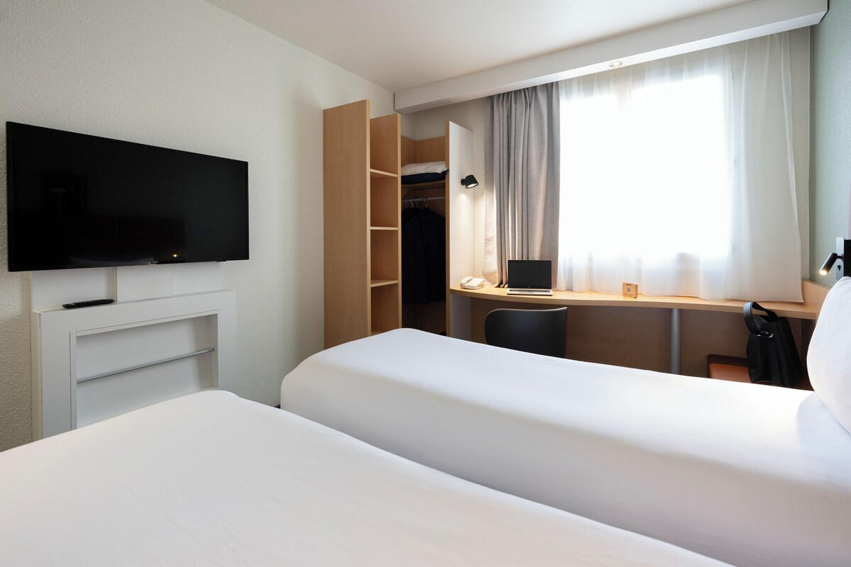 Habitación de hotel con cama, escritorio con computadora portátil, televisor, armario y ventana con cortinas.