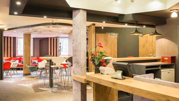 Un espace de bureau moderne doté d’un long comptoir d’accueil en bois, de plusieurs coins salon avec des chaises rouges et blanches, et d’une grande fenêtre laissant entrer la lumière naturelle.