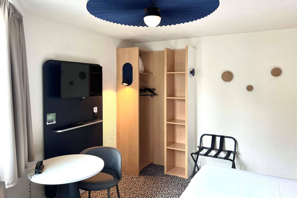 A compact hotel room with a bed, a small table, a chair, a wardrobe, a desk with a chair, a ceiling fan, and a wall-mounted TV.