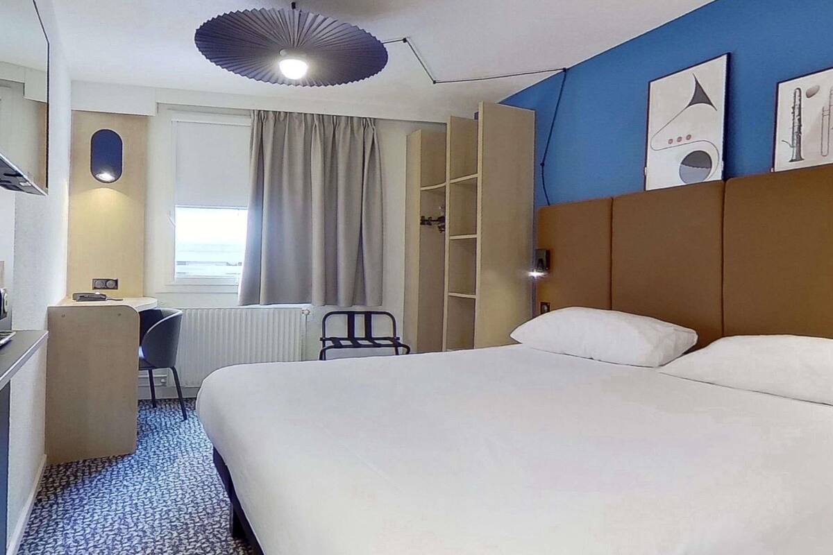 Ein Hotelzimmer mit Bett, Schreibtisch, Stuhl, Heizkörper, einem Fenster mit Vorhängen und einer Deckenleuchte.