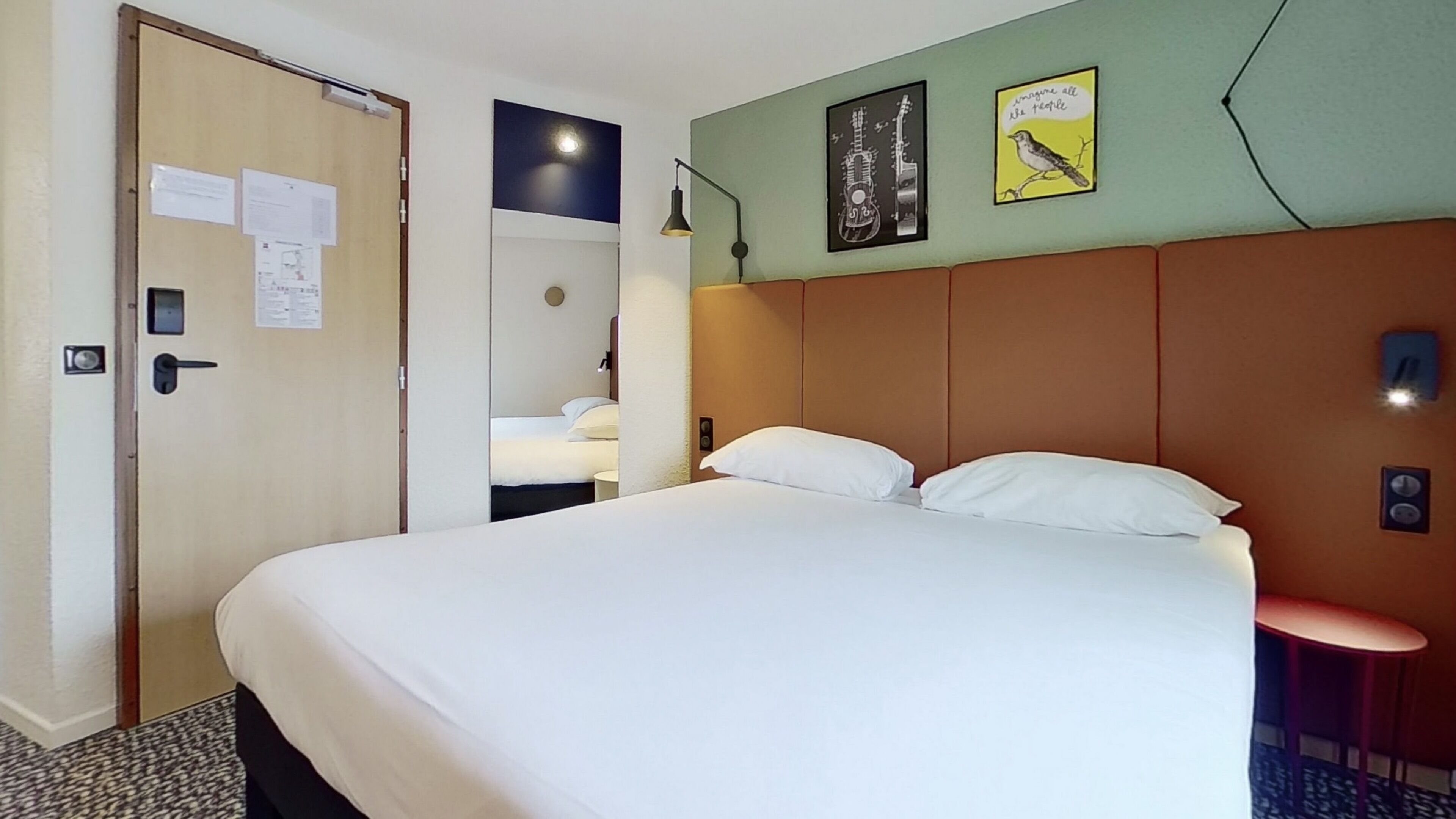 Habitación de hotel con una cama grande, una puerta con cerradura, una mesita y un cuadro con dos imágenes enmarcadas.