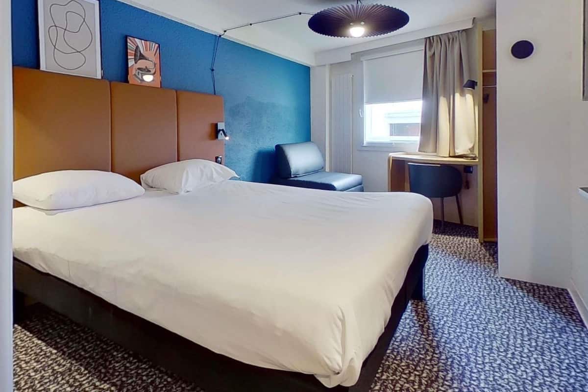 Ein Hotelzimmer mit einem großen Bett, einem Schreibtisch, einem Stuhl, einer Deckenleuchte und einem Vorhangfenster.