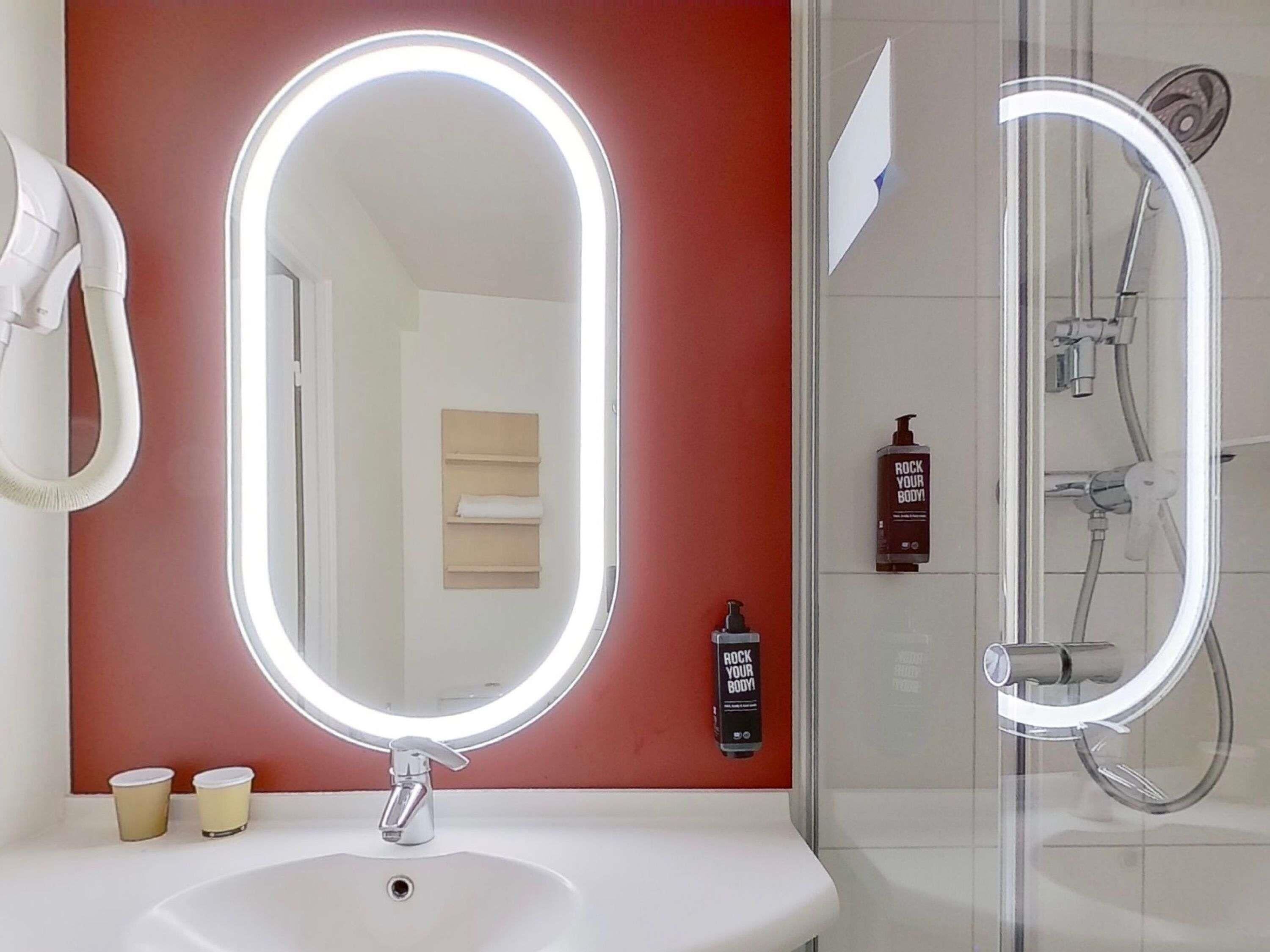 A bathroom with a white sink, two cup holders, a mirror with a light, a shower with a glass door, and a red accent wall.