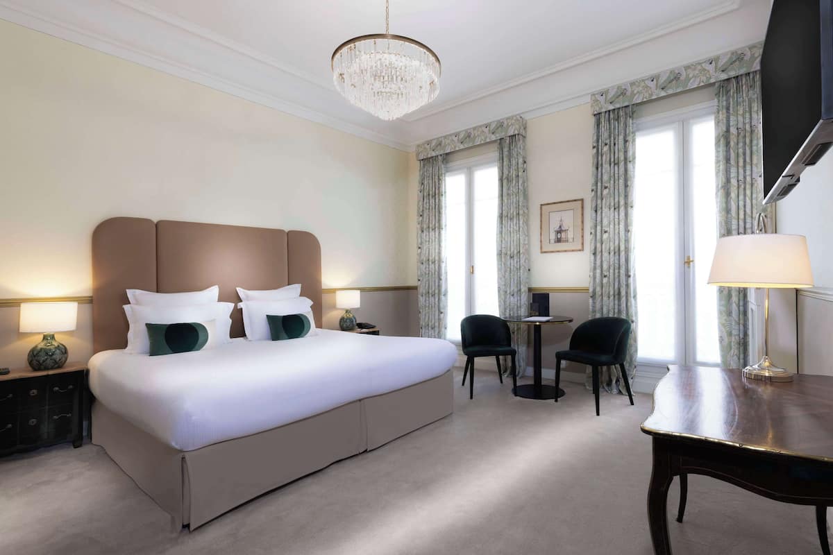 A hotel room with a large bed, two bedside tables with lamps, a desk, two chairs, a television, and a chandelier.