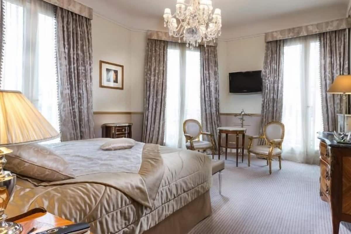 A hotel room with a bed, a desk, two chairs, a television, a chandelier, and a dresser.