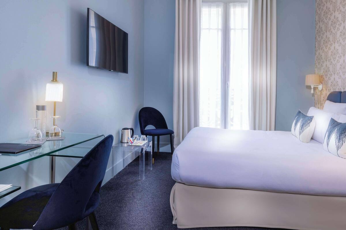 A hotel room with a bed, a desk with a chair, a television, and a window with curtains.