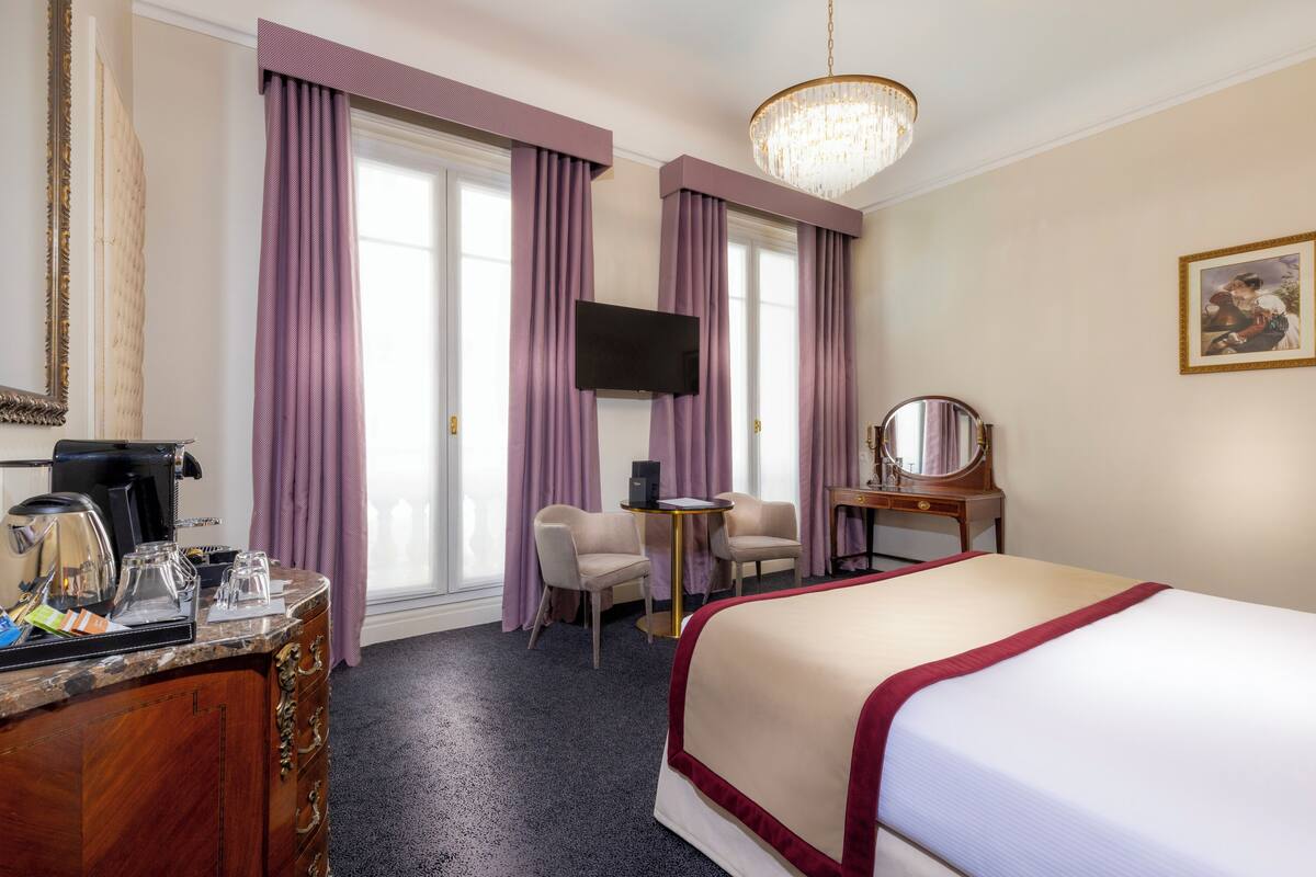 A hotel room with a bed, a desk with a chair, a television, a coffee maker, and a large window with purple curtains.