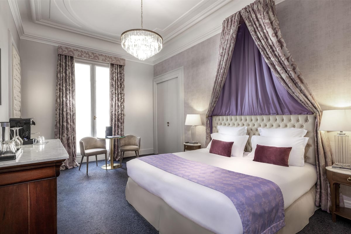 A hotel room with a large bed, a chandelier, a desk with a coffee maker, and a chair.
