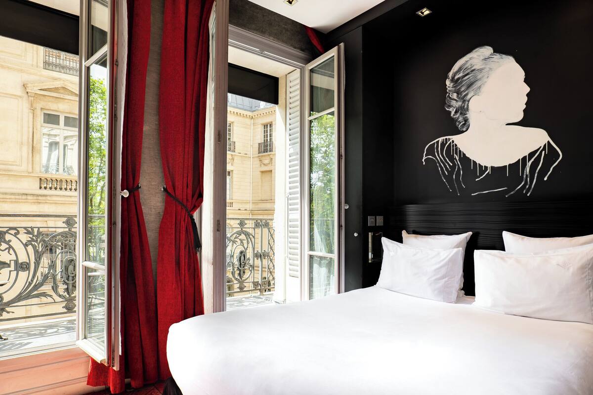 Ein Hotelzimmer mit einem großen Bett, einem Balkon mit roten Vorhängen und einer schwarzen Wand mit einer weißen Silhouette.