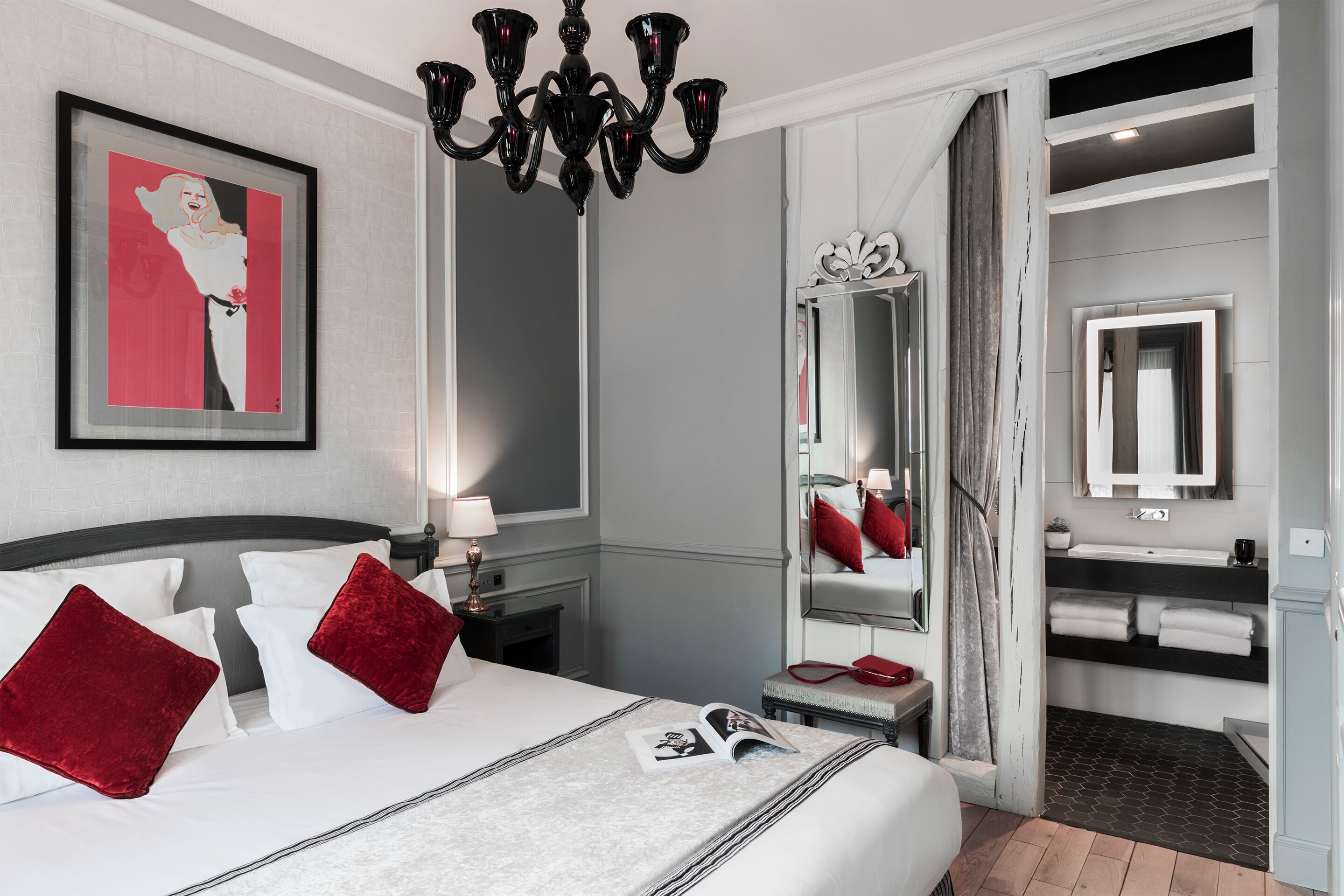 Une chambre d’hôtel avec un grand lit, un lustre, un tableau encadré, une coiffeuse avec un miroir et une salle de bain équipée d’un lavabo et d’un porte-serviettes.