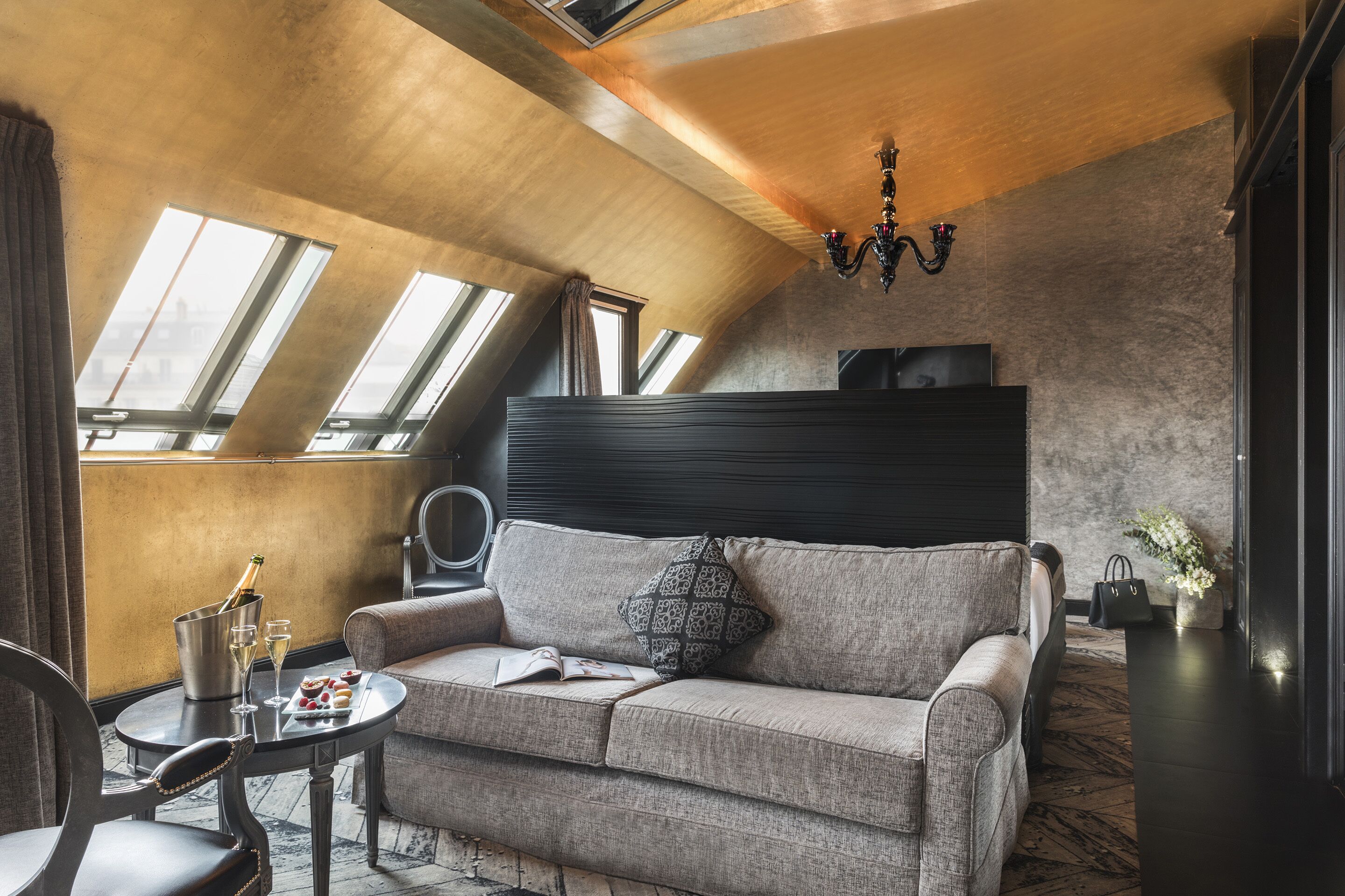 Um quarto moderno estilo loft com teto alto, claraboias, um sofá, uma pequena mesa com bebidas e um lustre decorativo.