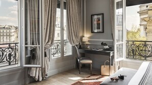 Een kamer met een bureau, stoel en bed. Gordijnen hangen voor de ramen, en er is uitzicht op gebouwen.