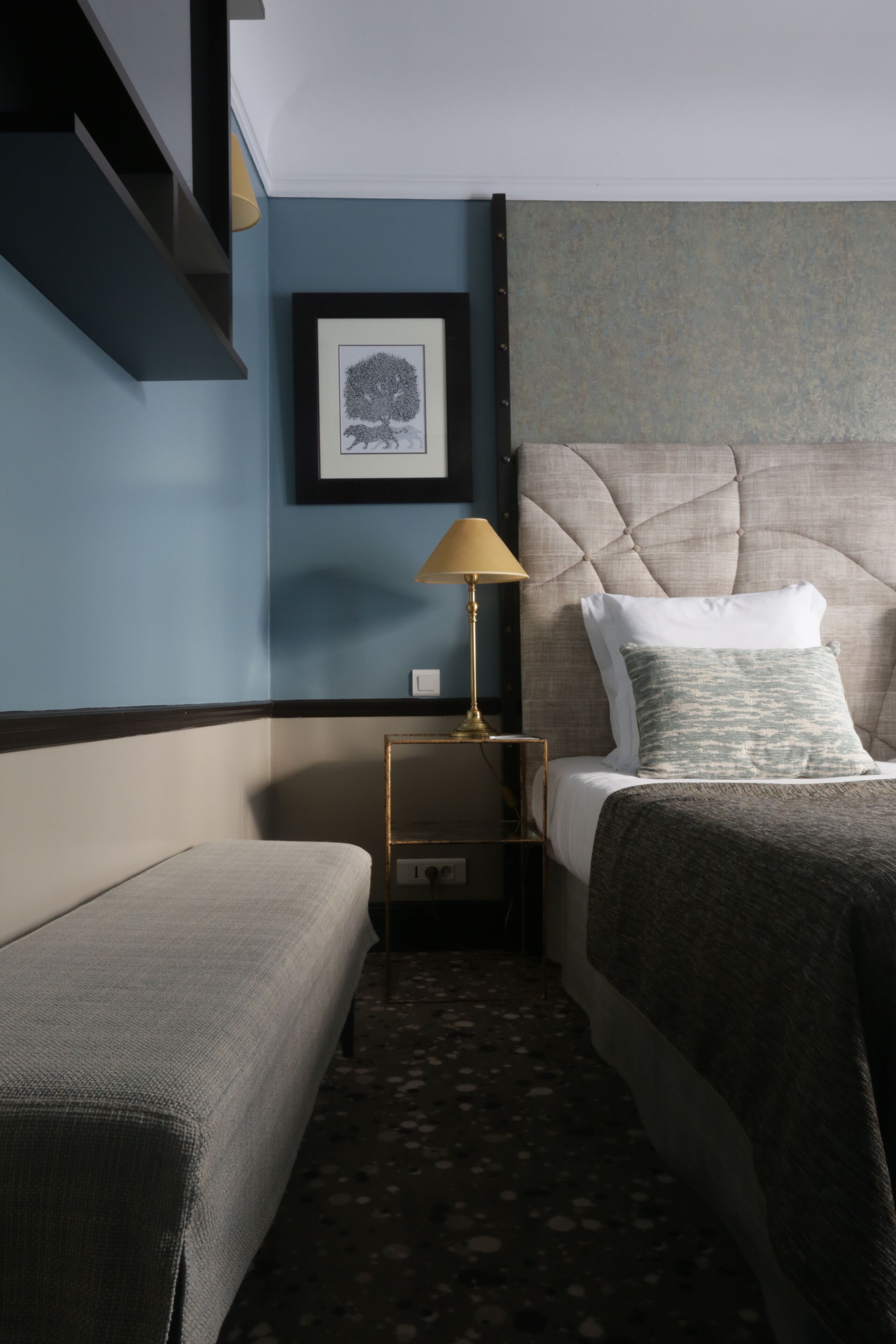 Et hotelværelse med en seng, et natbord med lampe, et billede i ramme på væggen og et tæppe på gulvet.