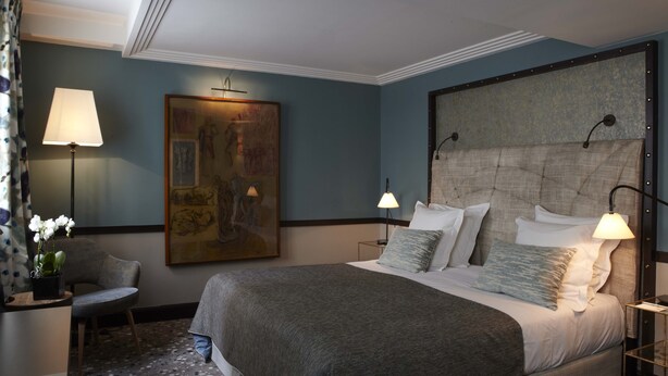 A hotel room with a large bed, two bedside lamps, a painting on the wall, a chair, and a small table with a plant. - Hotel Therese (Paris)