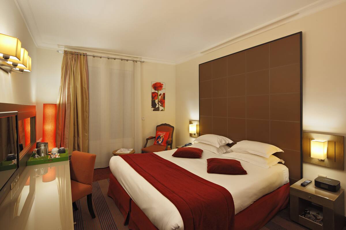 A hotel room with a large bed, a desk with a chair, a TV, and a nightstand with a lamp.