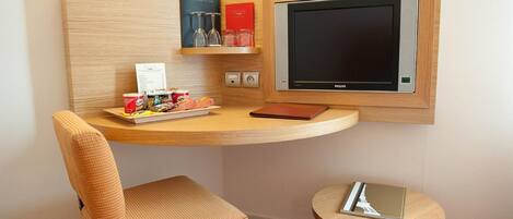 A compact hotel room with a wooden desk, a chair, a small round table, and a wall-mounted TV.