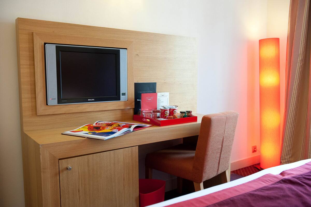 A hotel room with a wooden desk, a television, a red chair, and a bedside table with a magazine.
