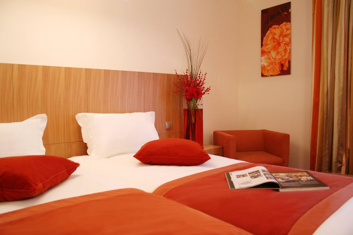 A hotel room with a wooden headboard, a bed with white and red bedding, a red chair, a vase with red flowers, and a framed picture on the wall.