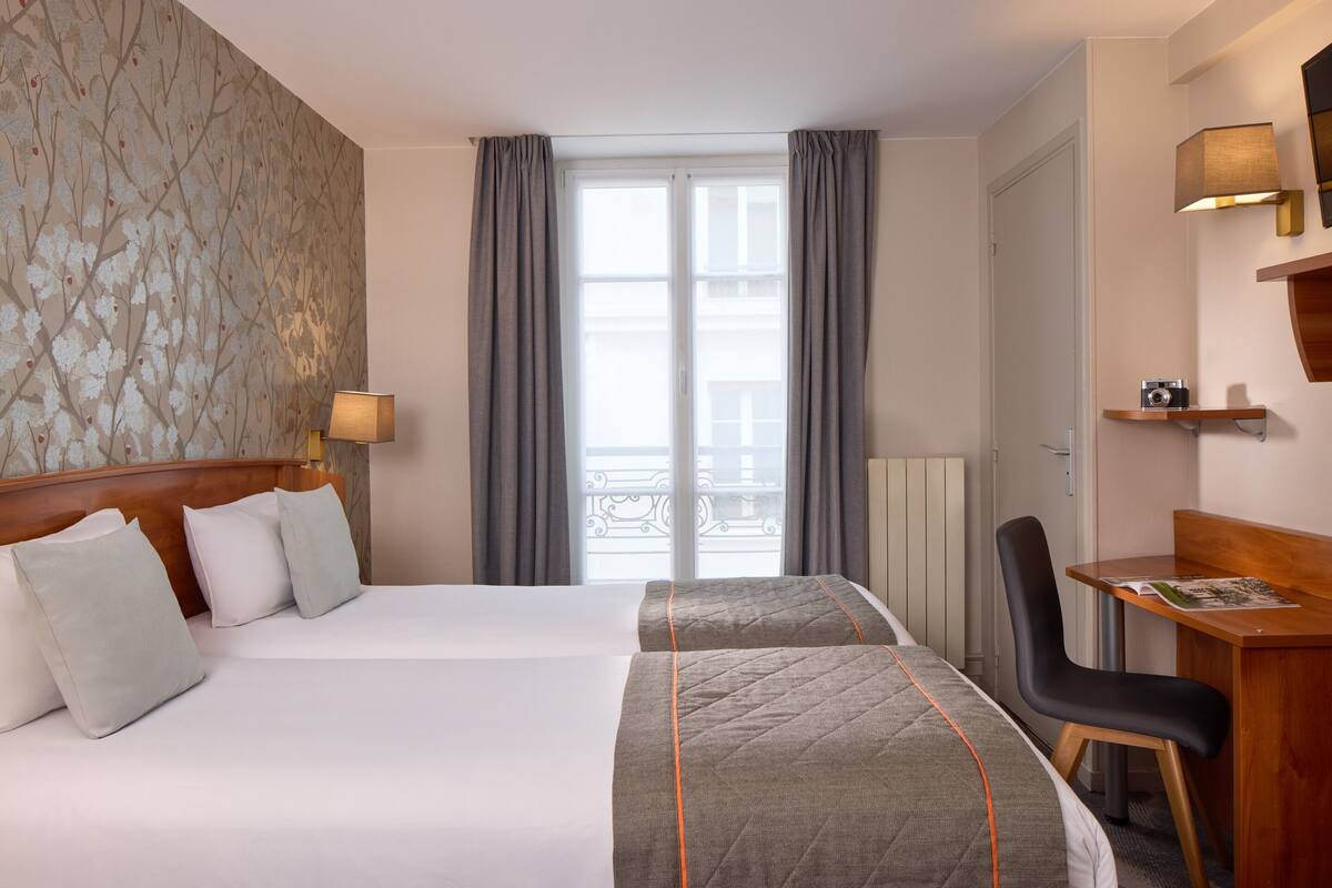 Une chambre d’hôtel avec deux lits, un bureau, une chaise, une lampe et une vue sur l’extérieur.