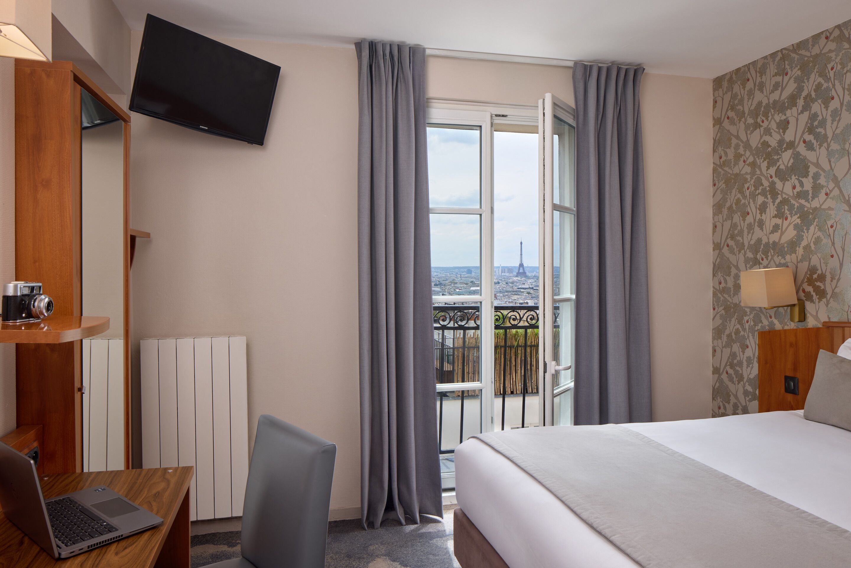 Ein Hotelzimmer mit einem Bett, einem Schreibtisch mit einem Laptop, einem Sessel, einem an der Wand montierten Fernseher und einem Balkon mit Blick auf den Eiffelturm.