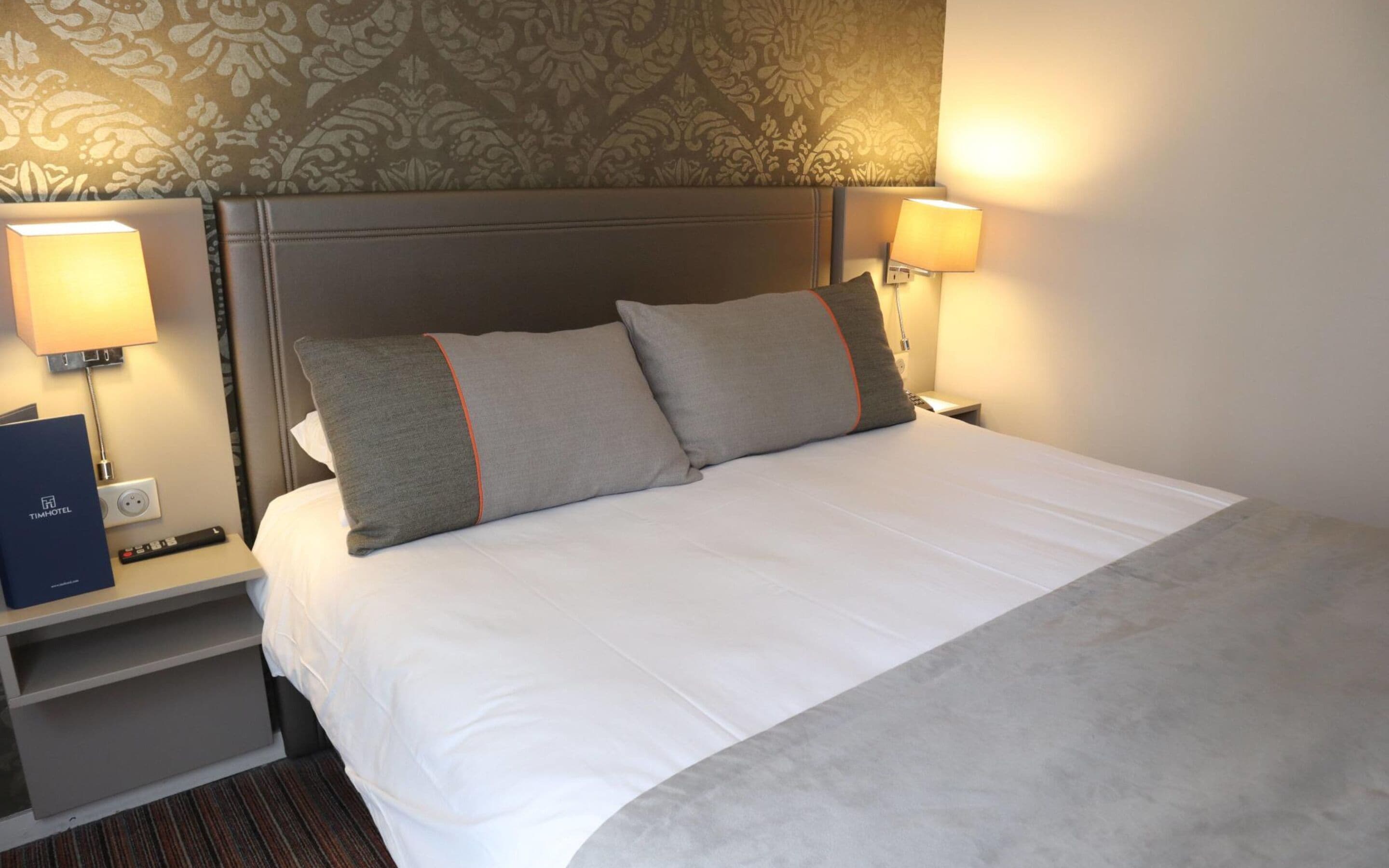 A hotel room with a neatly made bed, two bedside lamps, a remote control, and a TimHotel brochure on the nightstand.