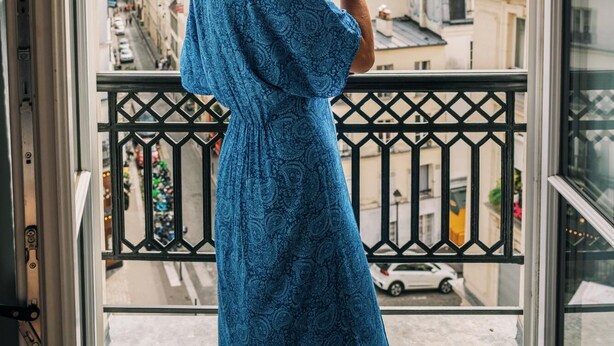 Eine Person in einem blauen Kleid steht an einem Fenster mit Blick auf eine Stadtlandschaft.