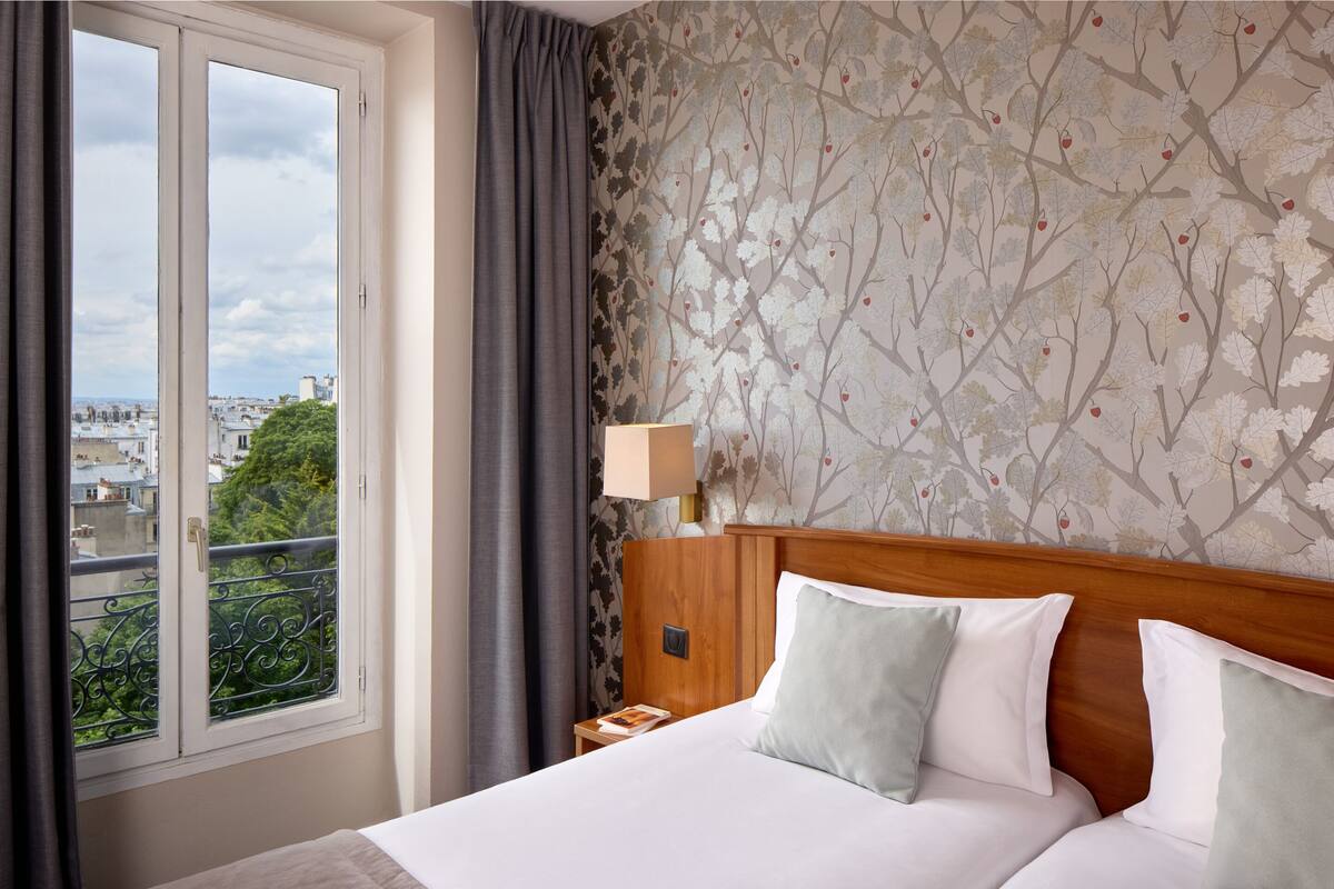 Une chambre d’hôtel dotée d’une grande fenêtre, d’un lit avec du linge de maison blanc, d’une tête de lit en bois et d’une table de chevet avec une lampe.