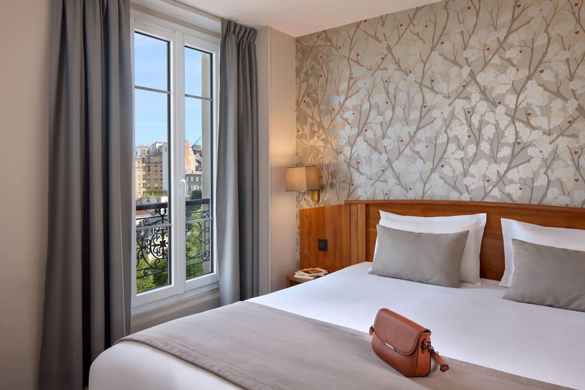 Une chambre d’hôtel avec un grand lit, une table de chevet, une lampe et une vue sur la ville, visible à travers une fenêtre munie de rideaux.