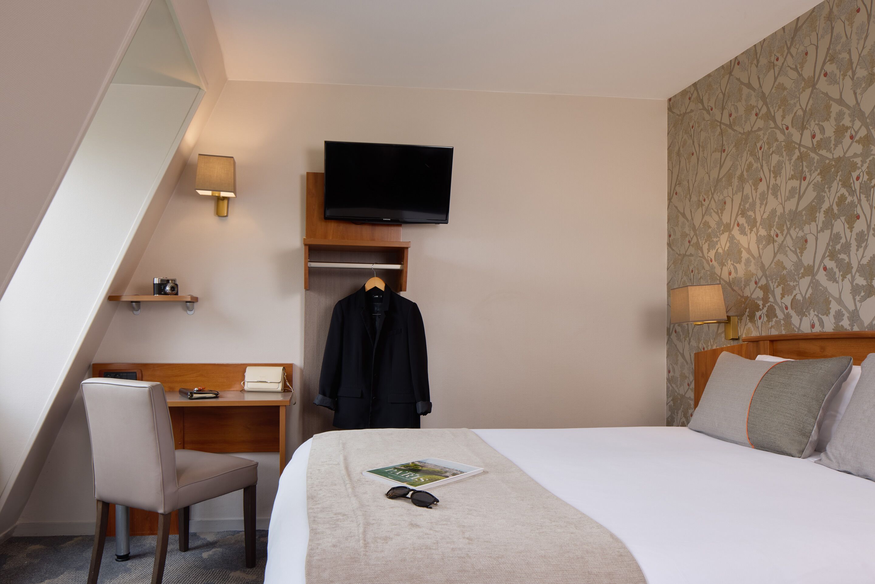 Habitación de hotel con cama, escritorio, silla, televisor montado en la pared y perchero.