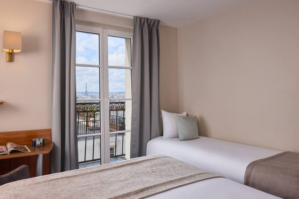 Une chambre d’hôtel avec un lit, un bureau doté d’une lampe et une vue sur la Tour Eiffel.