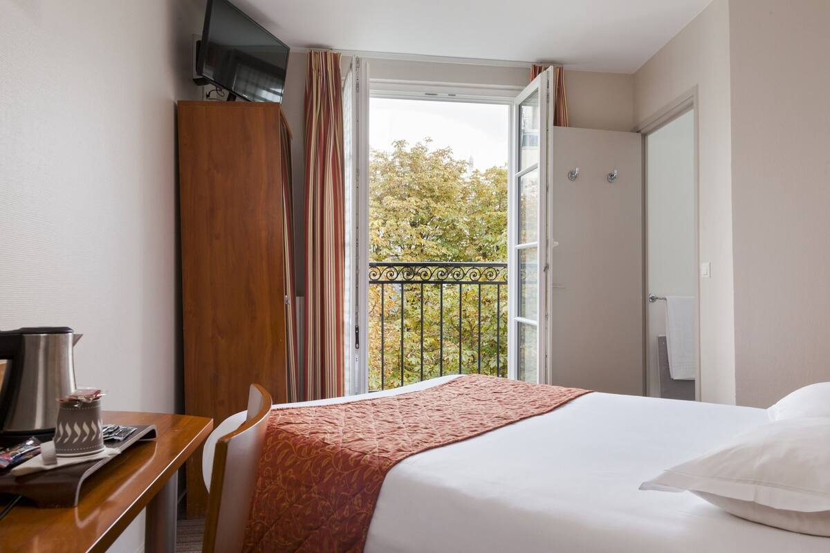 Une chambre d’hôtel comprenant un lit, un bureau avec une machine à café, un téléviseur posé sur un meuble et un balcon offrant une vue sur les arbres et les bâtiments.